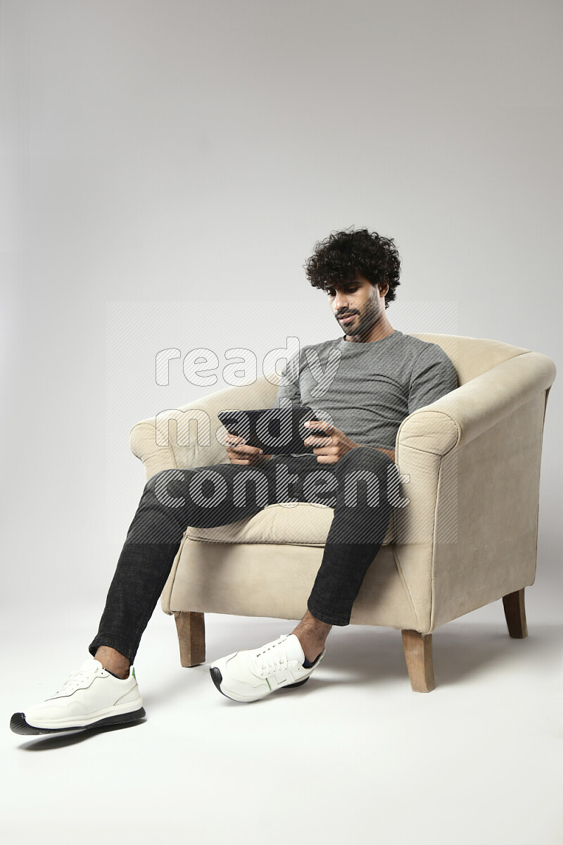 رجل يرتدي ملابس كاجوال يجلس علي كرسي و يلعب علي تابلت علي خلفية بيضاء
