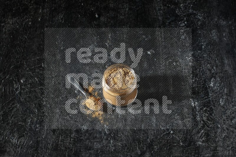 A glass jar and a metal spoon full of allspice powder on a textured black flooring
