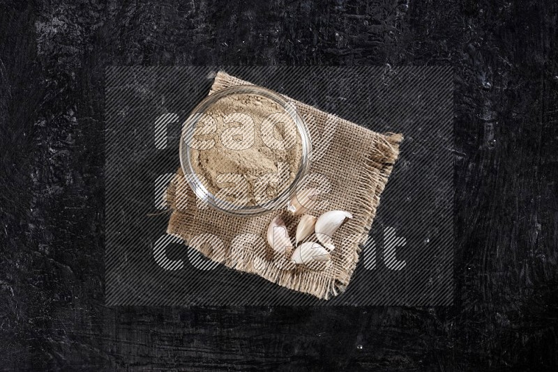 A glass bowl full of garlic powder on burlap fabric with garlic cloves on a textured black flooring in different angles