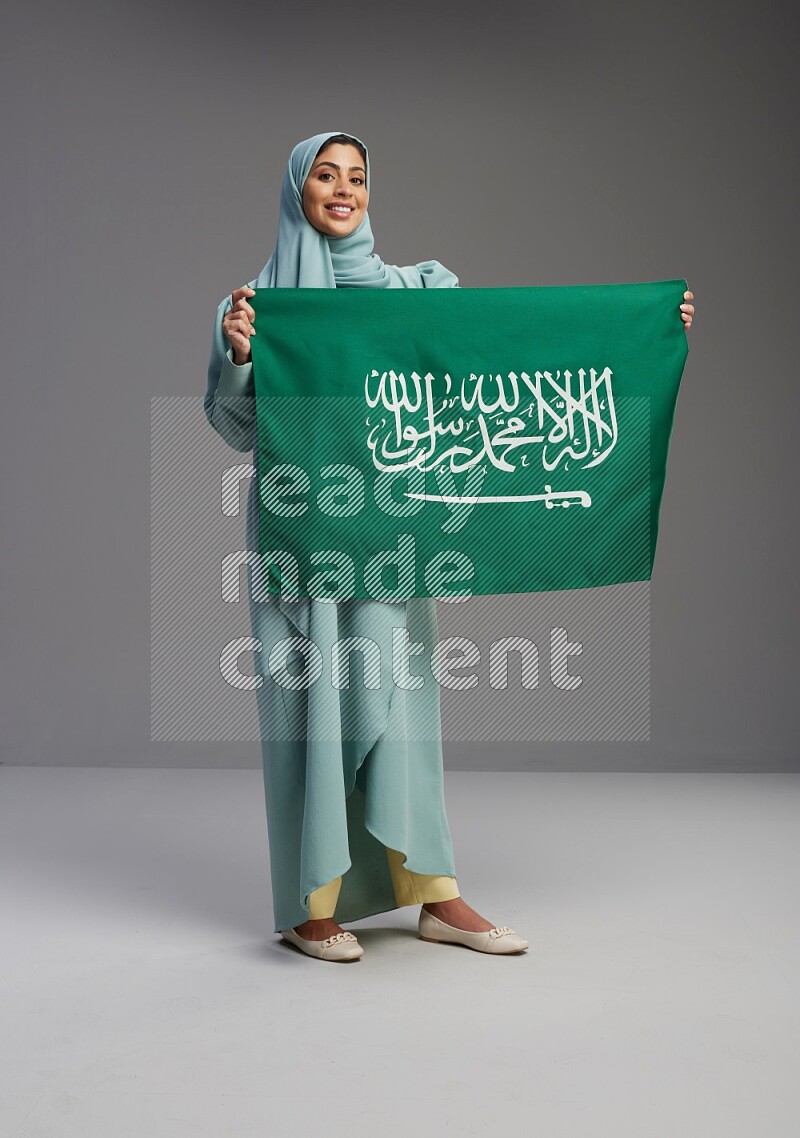 Saudi Woman wearing Abaya standing holding Saudi flag on Gray background