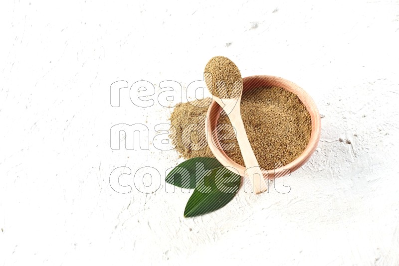 A wooden bowl and wooden spoon full of cumin powder on textured white flooring