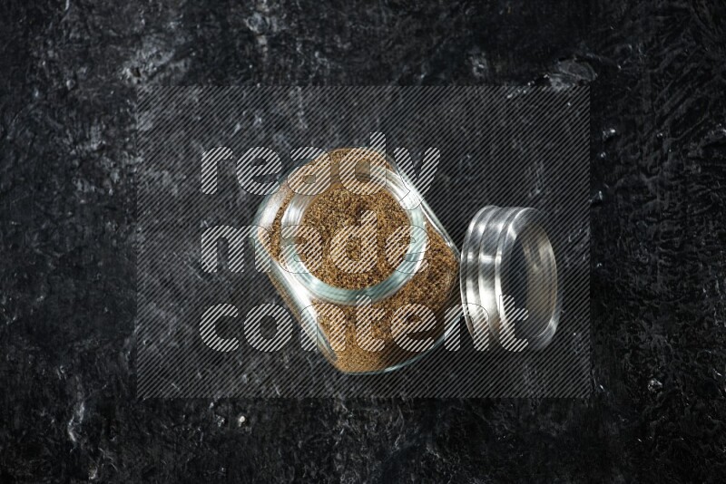 A glass spice jar full of cumin powder on a textured black flooring