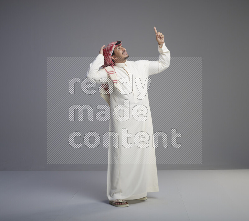 A Saudi man standing wearing thob and red shomag interacting with the camera on gray background