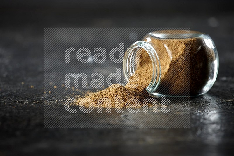 A flipped glass spice jar full of cumin powder and powder spilled out on a textured black flooring
