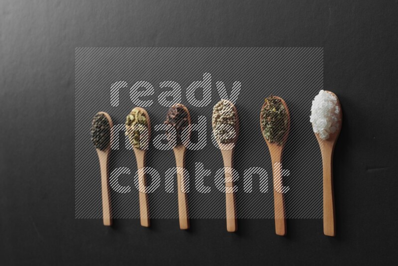 الفلفل الأبيض والقرنفل والهيل والملح والفلفل الأسود وريحان مجفف في معالق خشبية علي خلفية سوداء