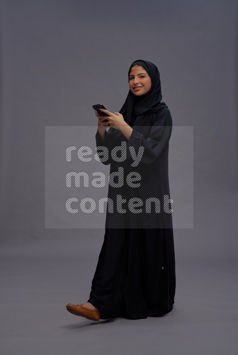 Saudi woman wearing Abaya standing texting on phone on gray background
