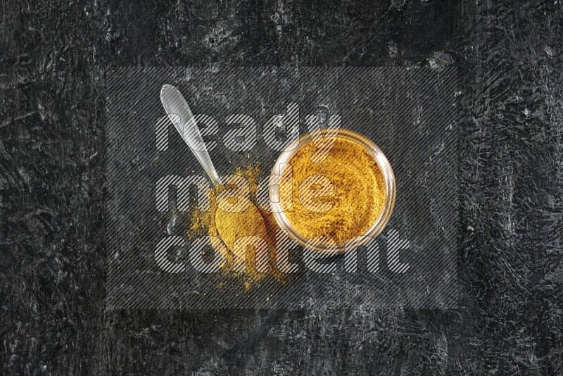 A glass jar and a metal spoon full of turmeric powder on a textured black flooring