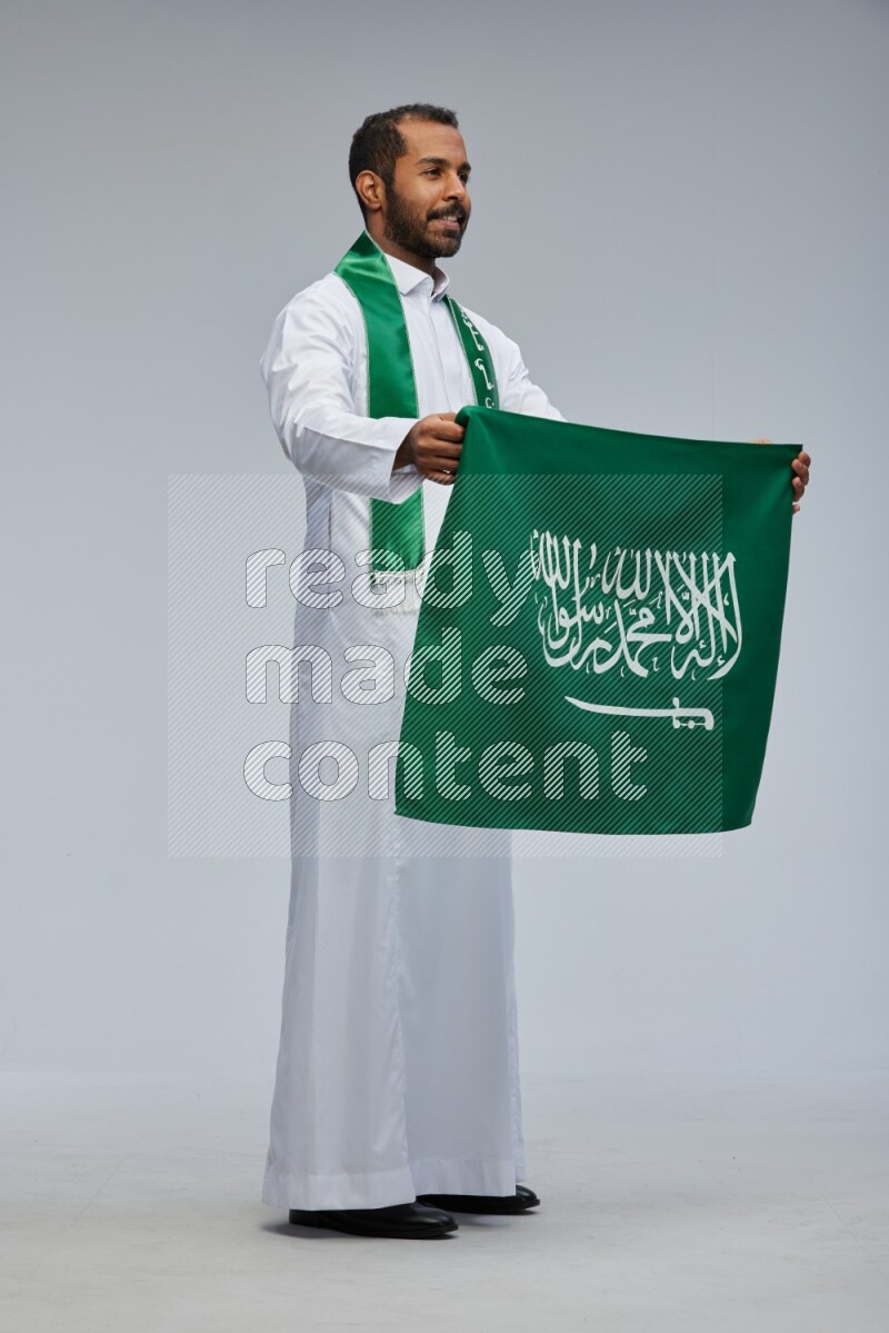 Saudi man wearing Thob standing holding Saudi flag on Gray background