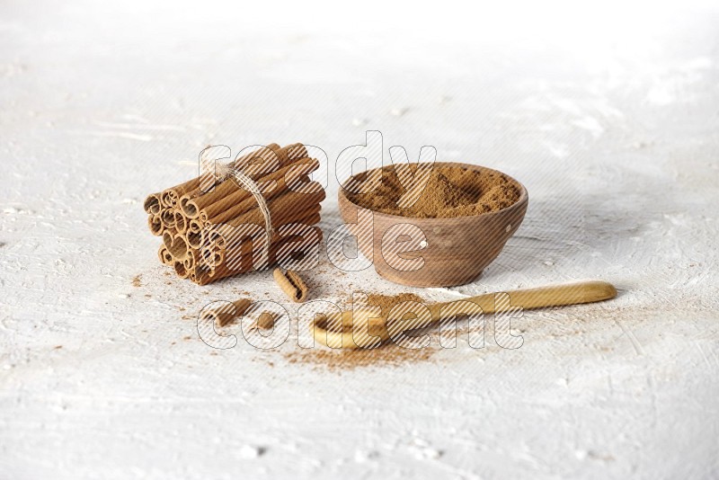 Cinnamon sticks stacked and bounded beside a wooden bowl full of cinnamon powder and a wooden spoon full of powder on white background