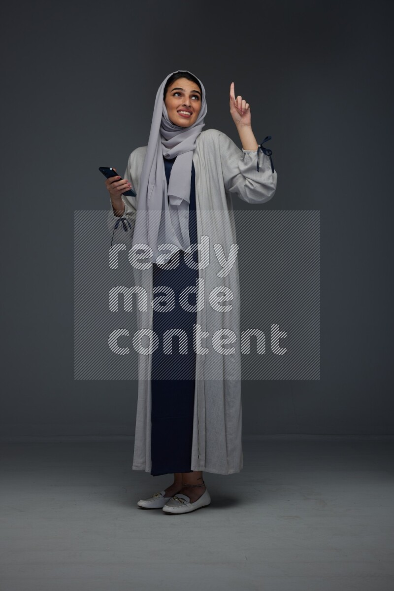 A Saudi woman wearing a light gray Abaya and head scarf standing and holding a phone on a grey background
