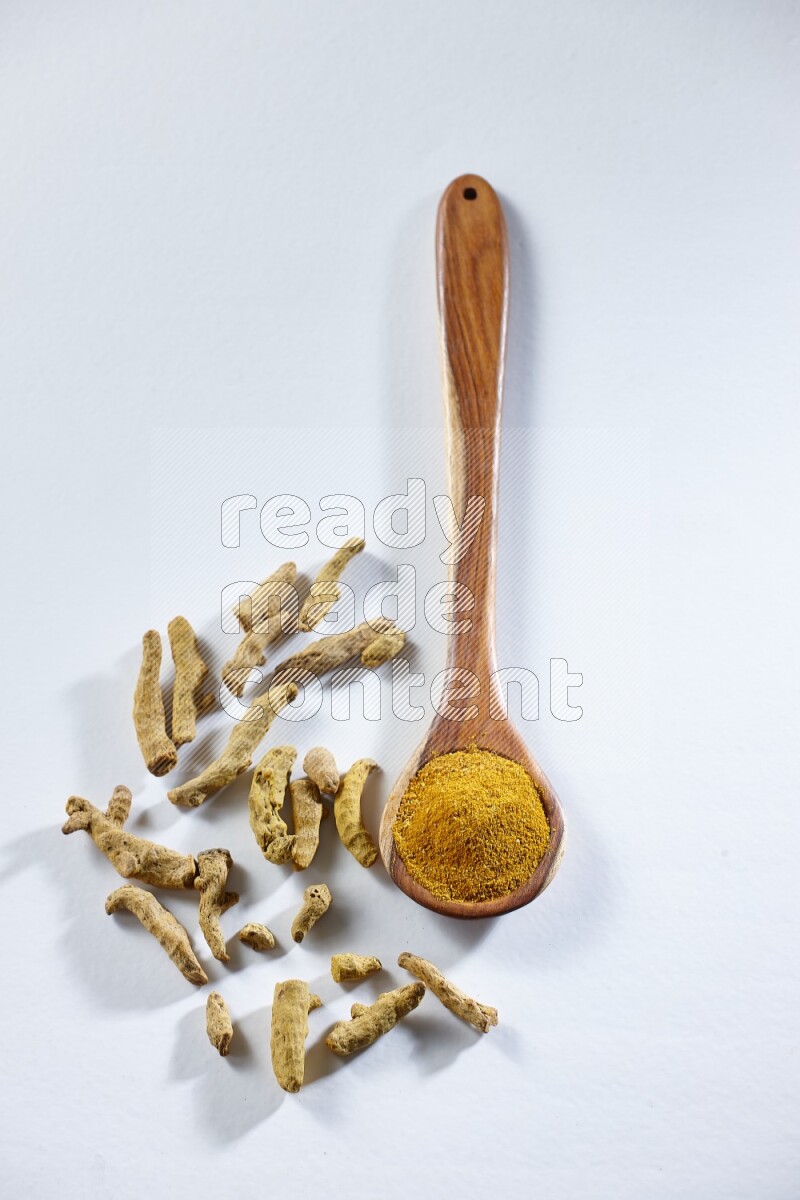 A wooden ladle full of turmeric powder and dried turmeric fingers beside it on white flooring