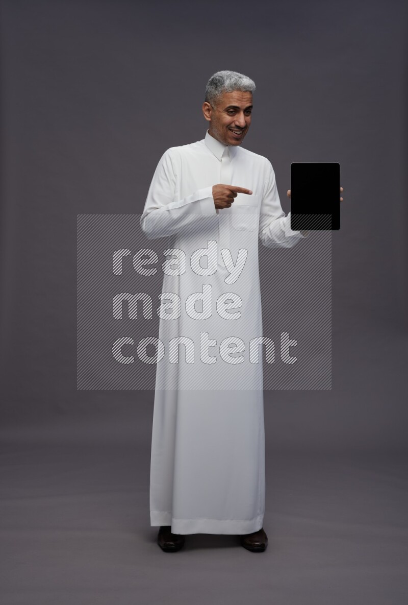 Saudi man wearing thob standing showing tablet to camera on gray background