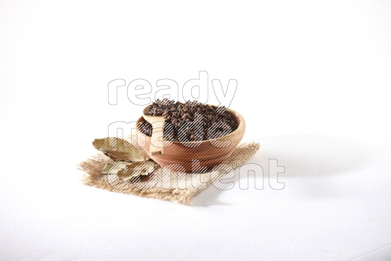 A wooden bowl and a wooden spoon full of cloves on a piece of burlap on a white flooring