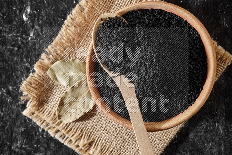 A wooden bowl full of black seeds with wooden spoon full of the seeds on it on a burlap fabric on a textured black flooring