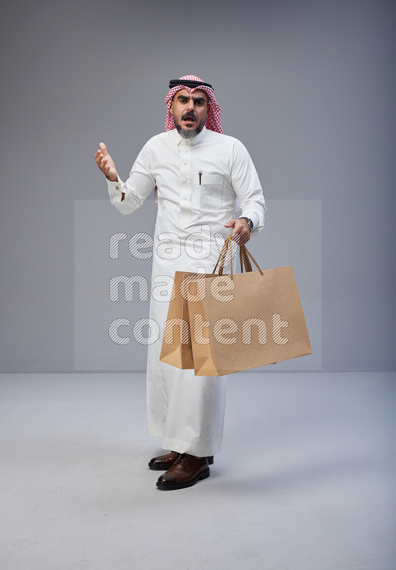 Saudi man Wearing Thob and red Shomag standing holding shopping bag on Gray background