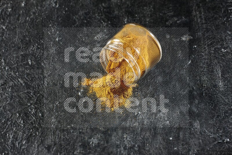 A flipped glass jar full of turmeric powder and powder spilled out of it on a textured black flooring