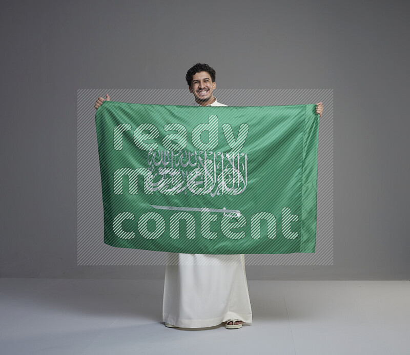 A saudi man standing wearing thob holding big saudi flag on gray background