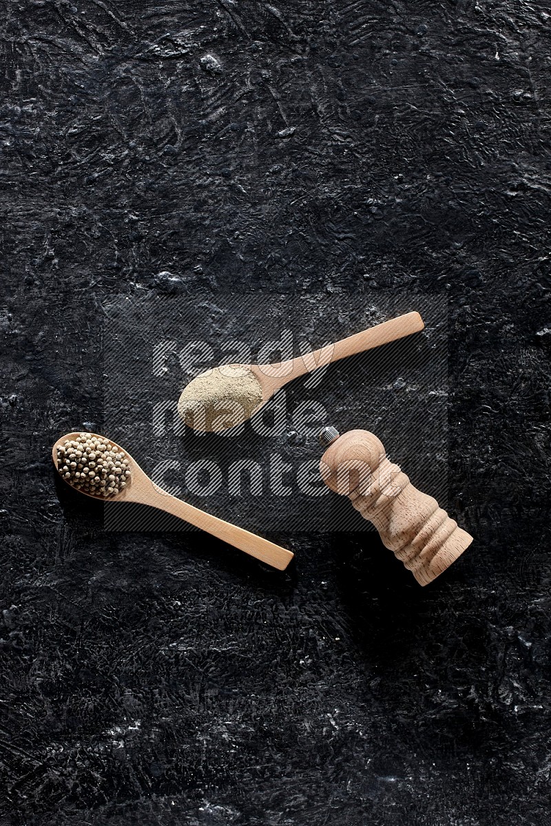 ملعقتان خشبيتان، واحدة ممتلئة ببودرة الفلفل الأبيض والأخرى بحبوب الفلفل ومطحنة خشبية على أرضية سوداء