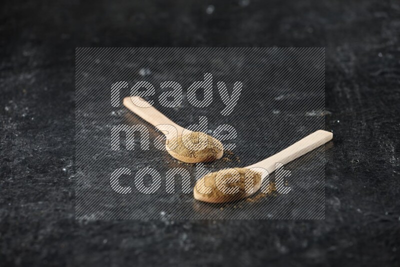 2 wooden spoons full of cumin powder on textured black flooring