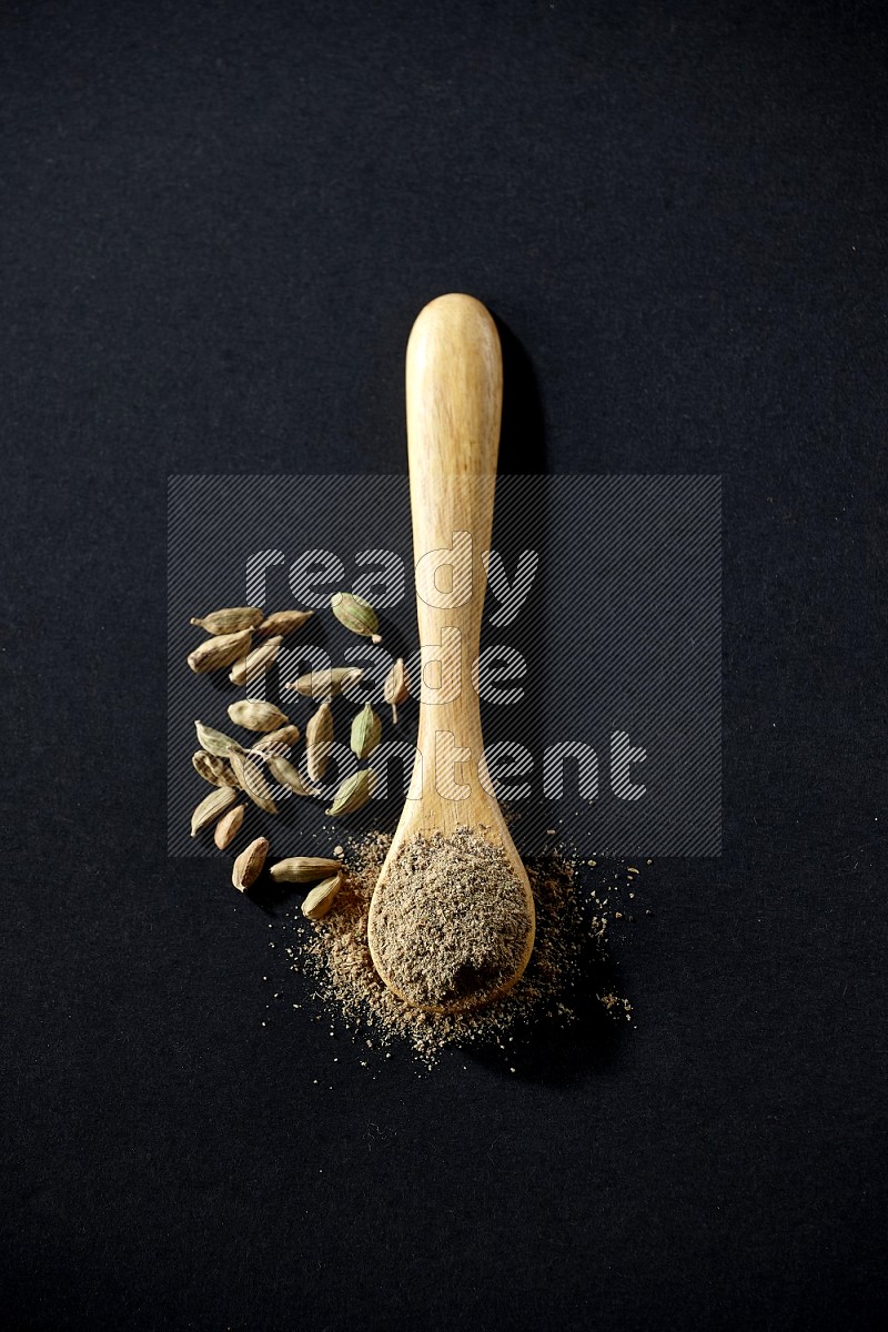 A wooden spoon full of cardamom powder and cardamom seeds beside it on black flooring