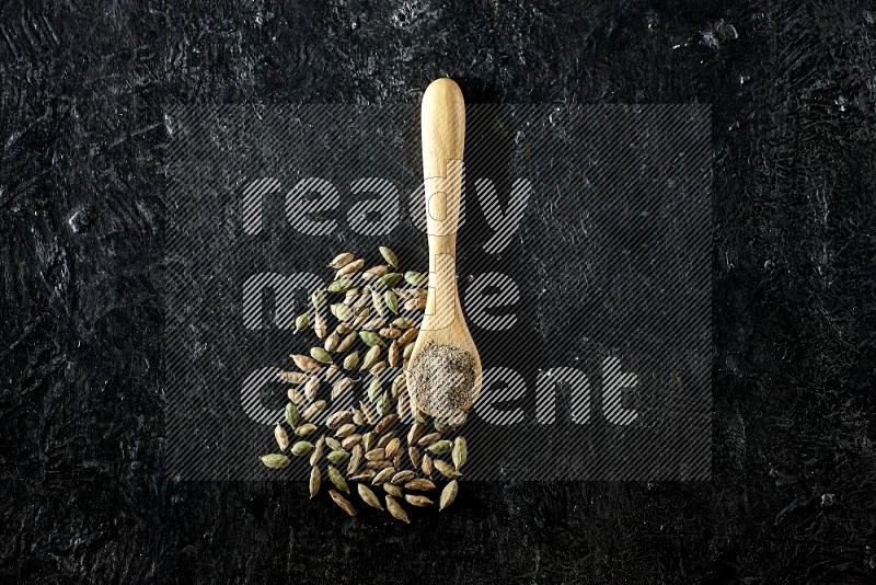 A wooden spoon full of cardamom powder and cardamom seeds next of it on textured black flooring