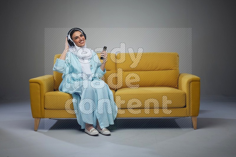 A Saudi woman wearing a light blue Abaya and a white head scarf sitting on a yellow sofa and holding an electronic while wearing headphone eye level on a grey background