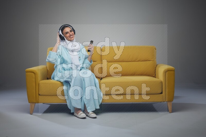 A Saudi woman wearing a light blue Abaya and a white head scarf sitting on a yellow sofa and holding an electronic while wearing headphone eye level on a grey background