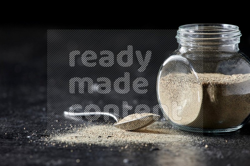 Herbal glass jar and metal spoon full of white pepper powder on textured black flooring