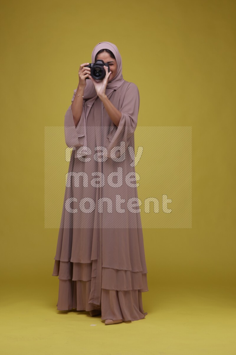 A woman Holding a Camera on a Yellow Background wearing Brown Abaya with Hijab