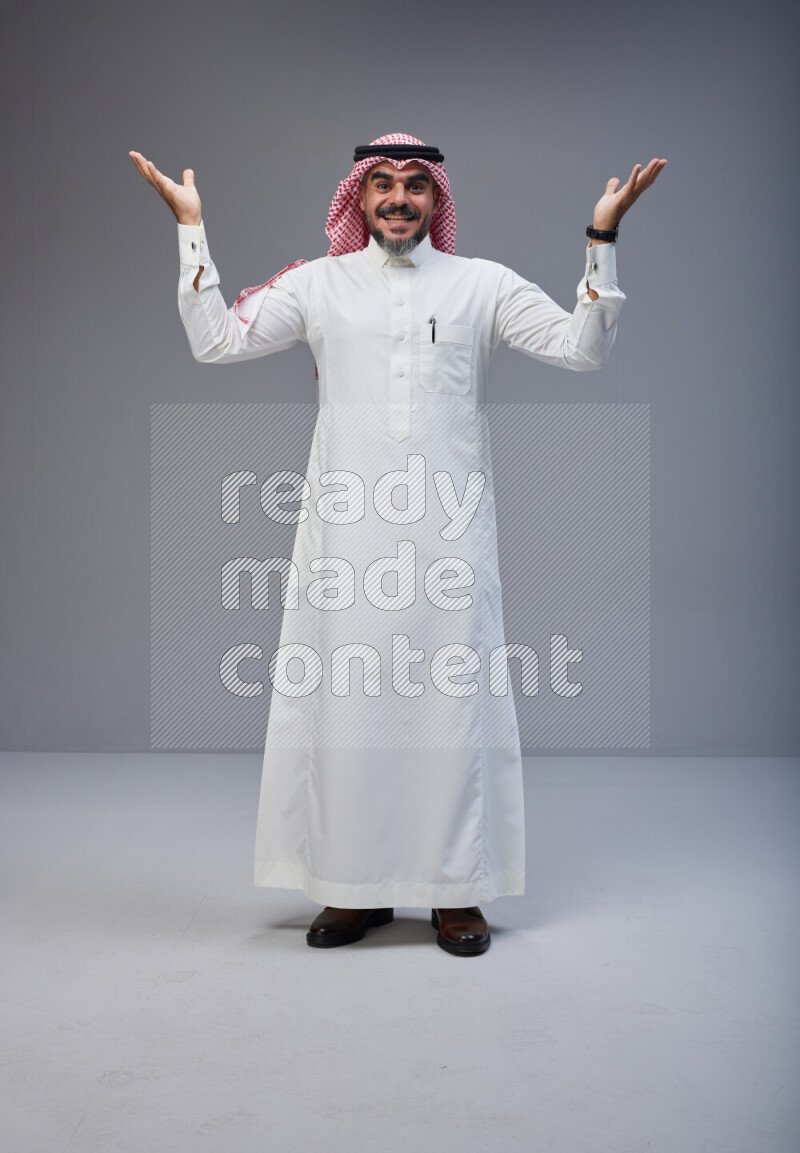 Saudi man Wearing Thob and red white Shomag standing interacting with the camera on Gray background