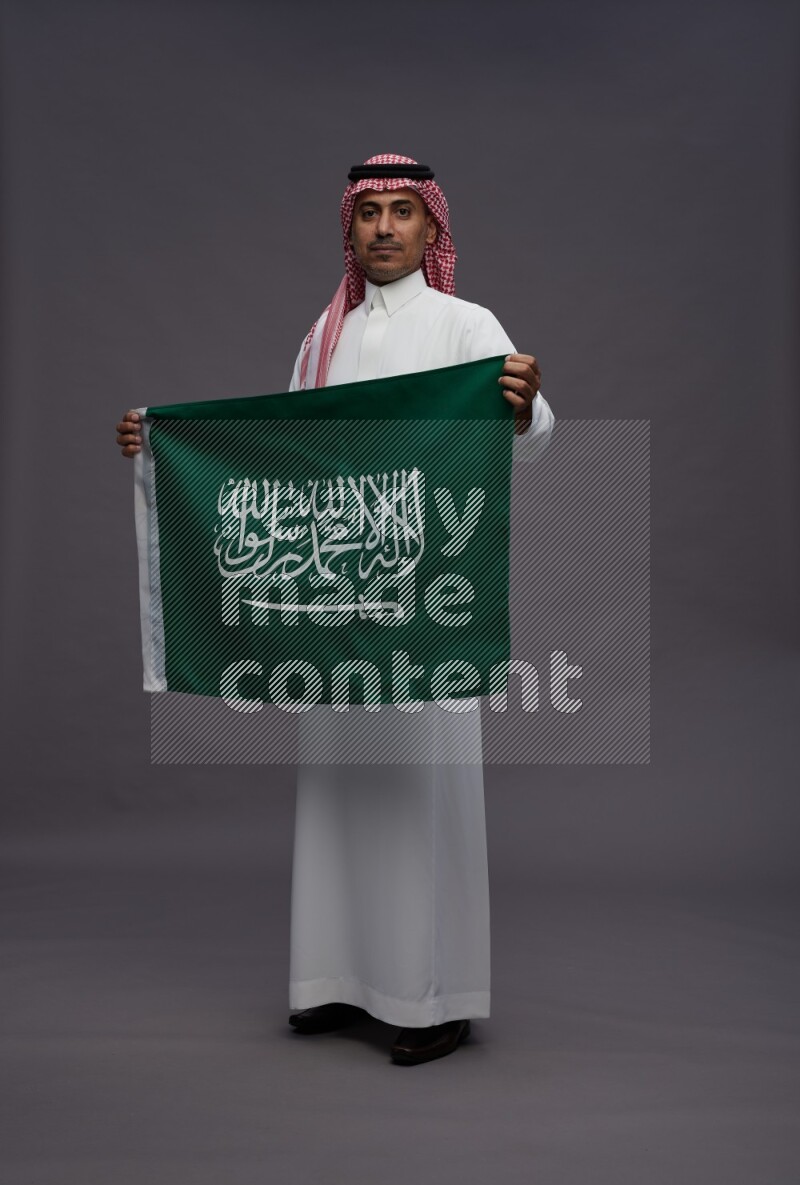 Saudi man wearing thob and shomag standing holding Saudi flag on gray background