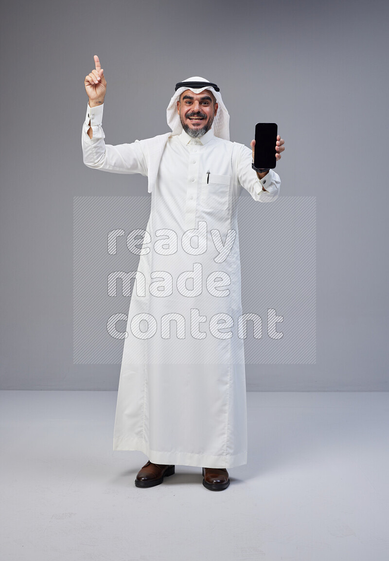 Saudi man Wearing Thob and white Shomag standing showing phone to camera on Gray background