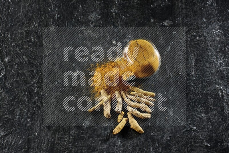 A flipped glass spice jar full of turmeric powder and powder spilled out of it with dried whole fingers on textured black flooring