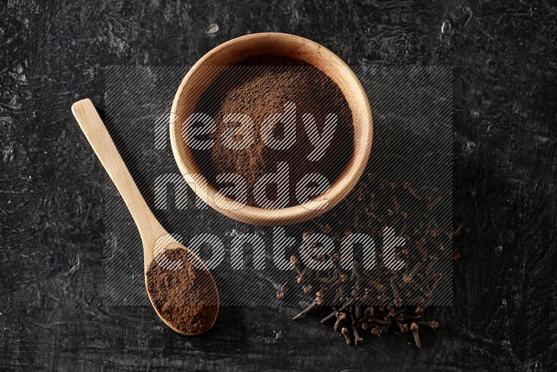 A wooden bowl and a wooden spoon full of cloves powder with spreaded cloves on a textured black flooring