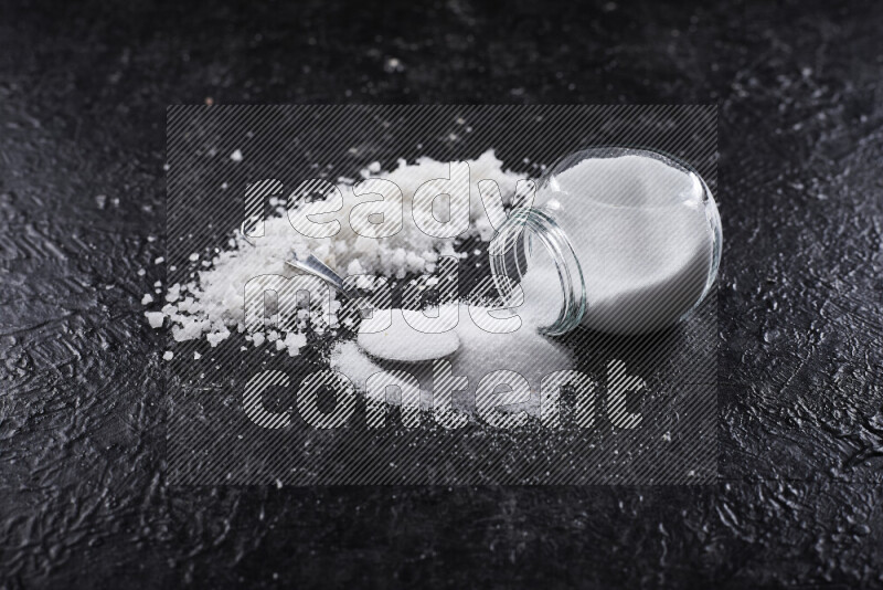 A glass jar full of table salt with some sea salt crystals beside it on a black background