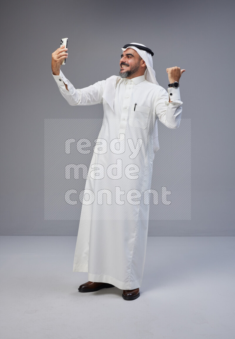 Saudi man Wearing Thob and white Shomag standing taking selfie on Gray background