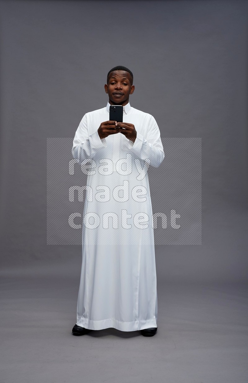 Saudi man wearing thob standing texting on phone on gray background