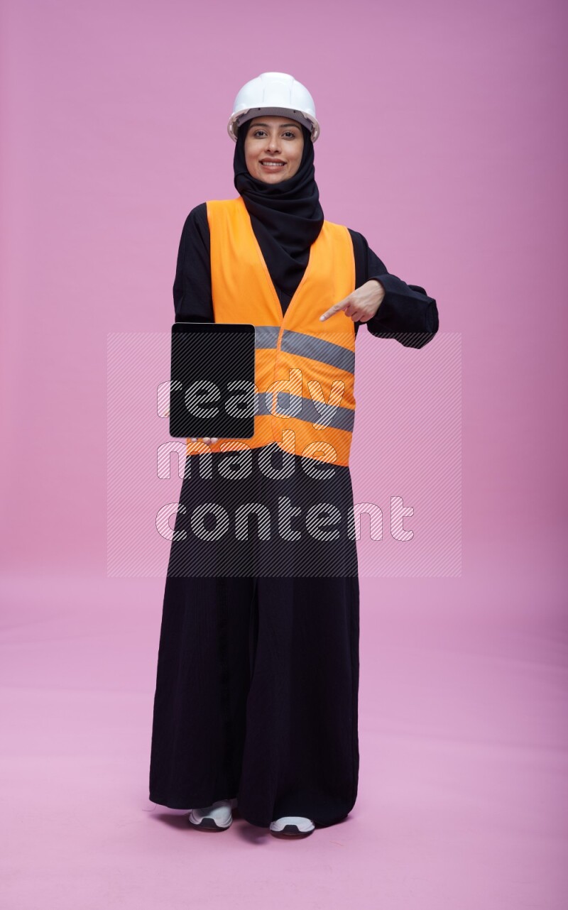 Saudi woman wearing Abaya with engineer vest and helmet standing showing tablet to camera on pink background