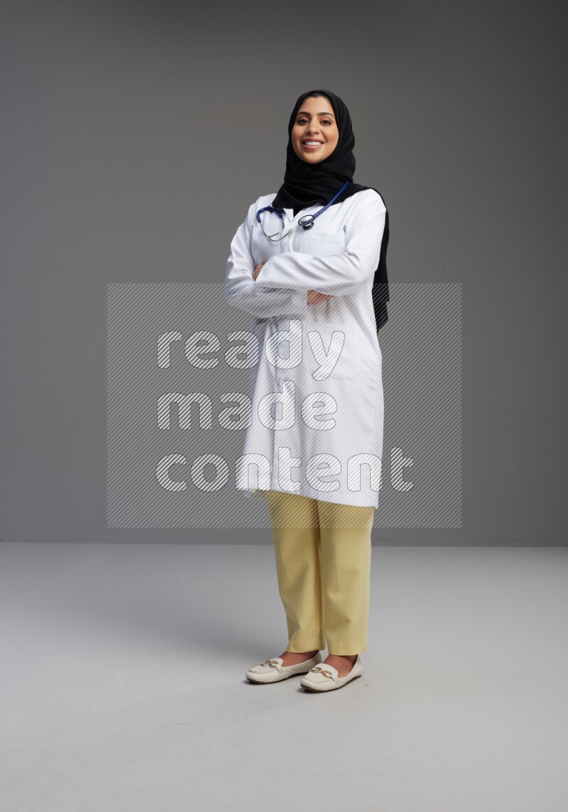 Saudi woman wearing lab coat with stethoscope standing with crossed arms on Gray background
