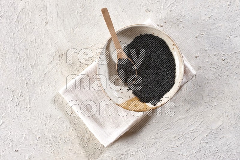 A multicolored pottery plate full of black seeds with a wooden spoon full of the seeds on a napkin on a textured white flooring in different angles