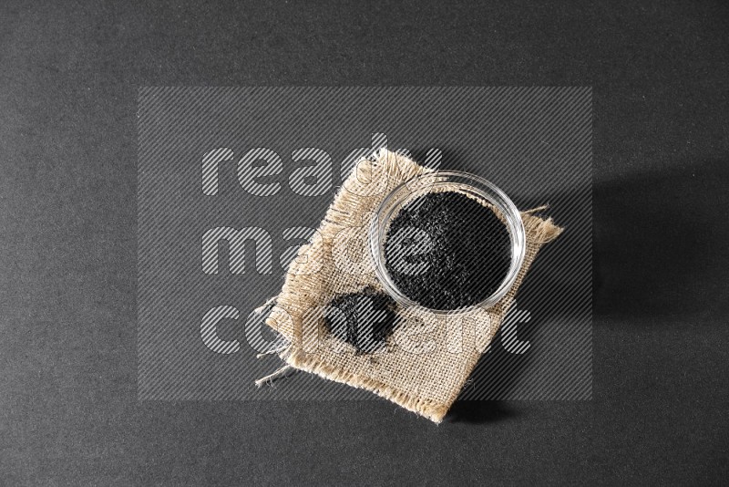 A glass bowl full of black seeds and seeds on burlap fabric on a black flooring