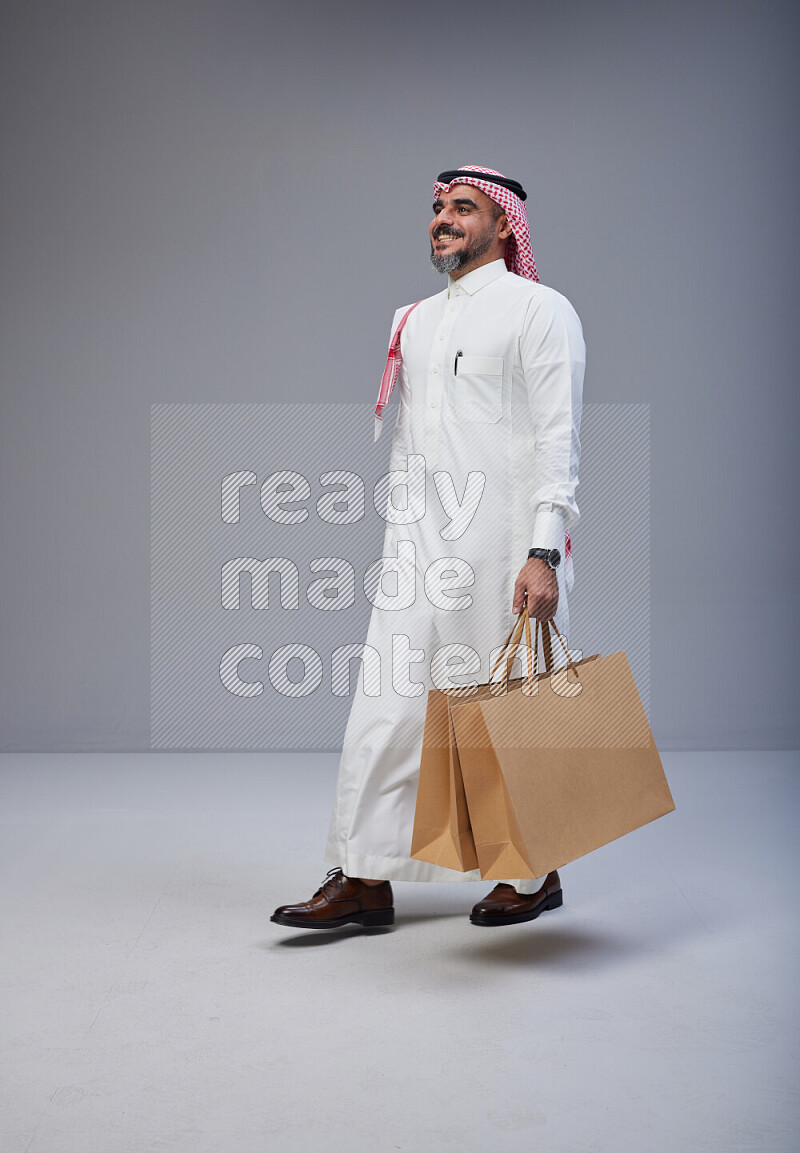 Saudi man Wearing Thob and red Shomag standing holding shopping bag on Gray background