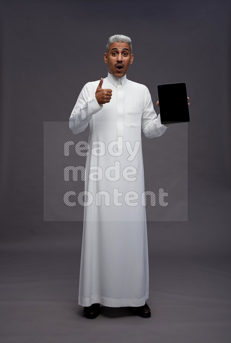 Saudi man wearing thob standing showing tablet to camera on gray background