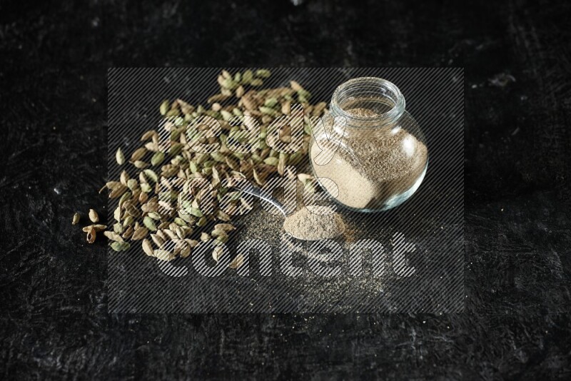 A glass spice jar and a metal spoon full of cardamom powder and cardamom seeds spreaded on textured black flooring