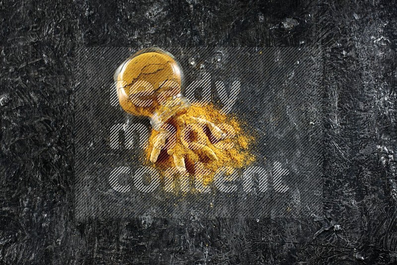 A flipped glass spice jar full of turmeric powder and powder spilled out of it with dried whole fingers on textured black flooring