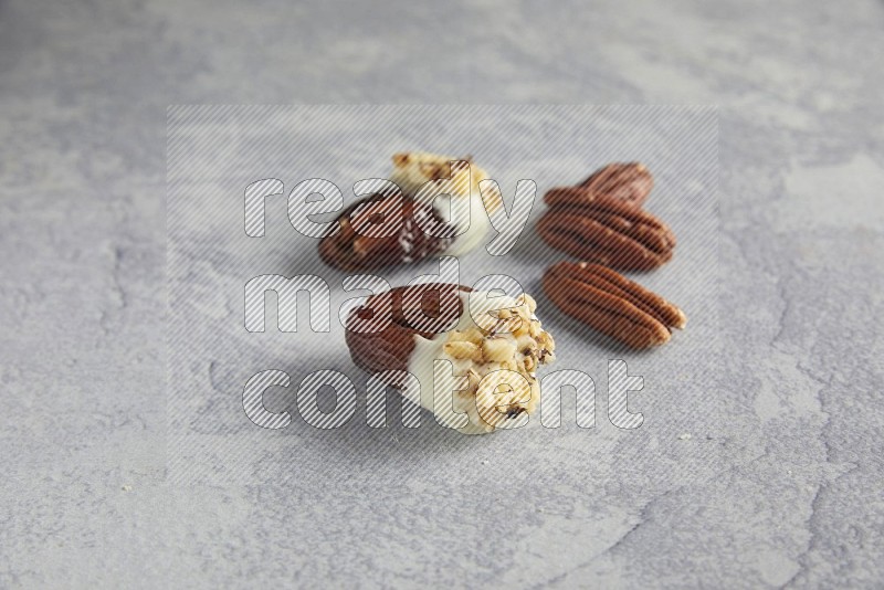 Two pecan stuffed dates covered with white chocolate and chopped walnuts with unroasted pecans on alight grey background