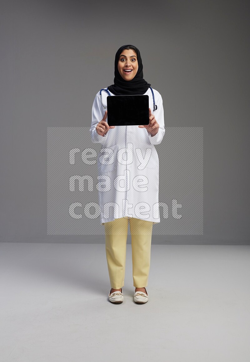 Saudi woman wearing lab coat with stethoscope standing showing tablet to camera with sign in the back on Gray background