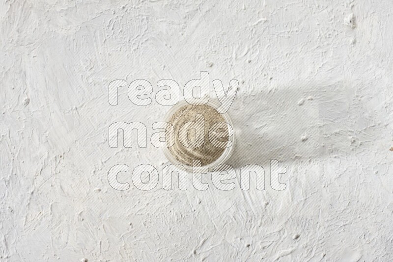 A glass jar full of white pepper powder on textured white flooring