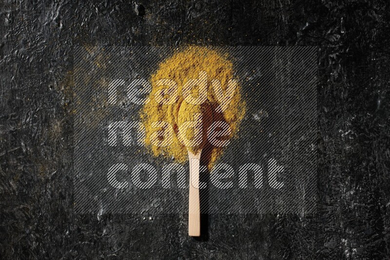 A wooden spoon full of turmeric powder on textured black background