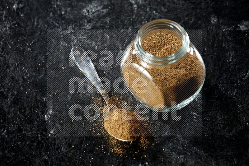 A glass spice jar and a metal spoon full of cumin powder on a textured black flooring